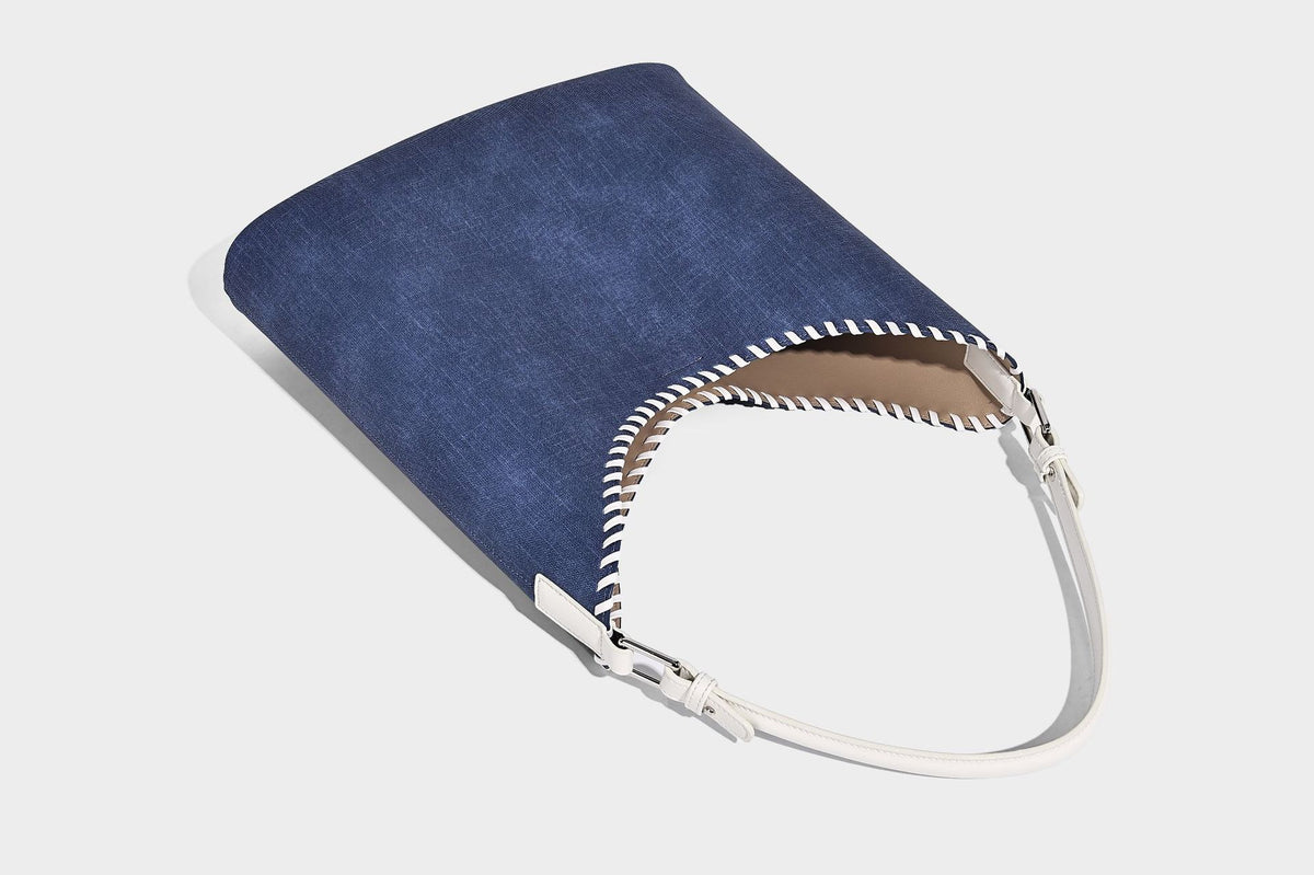Denim-blue hobo tote with white leather-look shoulder strap, contrast whipstitch trim along curved open top, and silver-tone