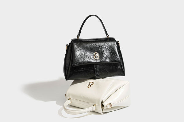 Structured black satchel bag with glossy crinkled leather-look finish, gold-tone emblem flap closure, slim top handle, and