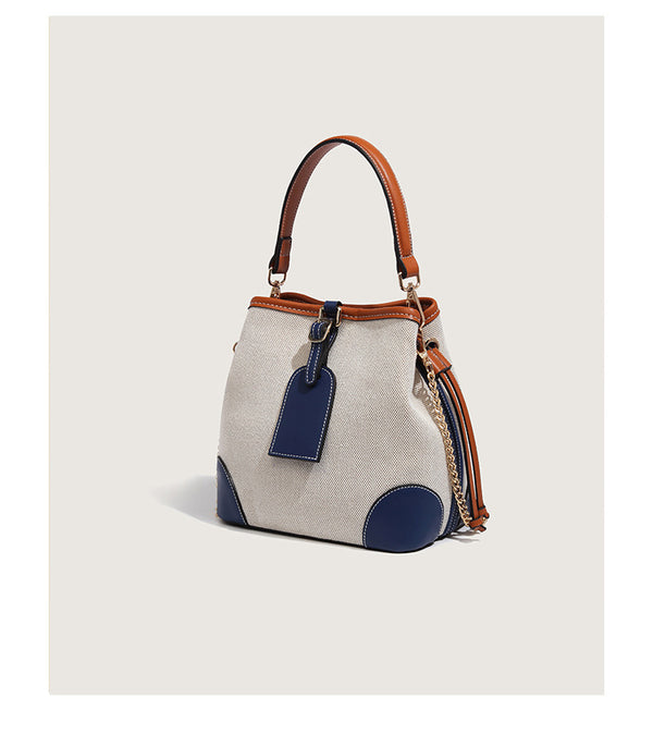 Cream woven-texture canvas bucket bag with navy accent panels, tan leather-look trim, gold-tone chain strap, and tassel