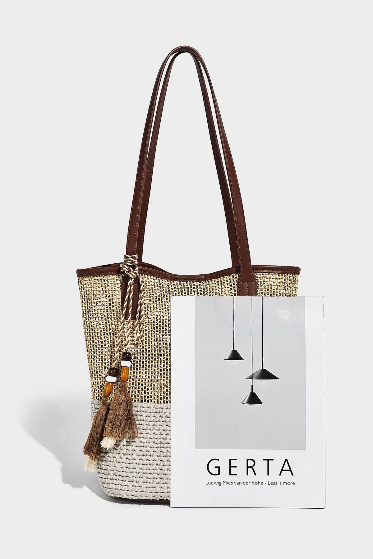 Woven straw-look tote bag in tan and cream with dark brown leather-look trim, long flat shoulder straps, and tassel bead