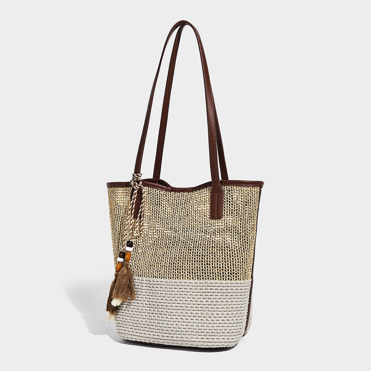 Woven straw-look tote bag in tan and cream with dark brown leather-look trim, long flat shoulder straps, and tassel bead