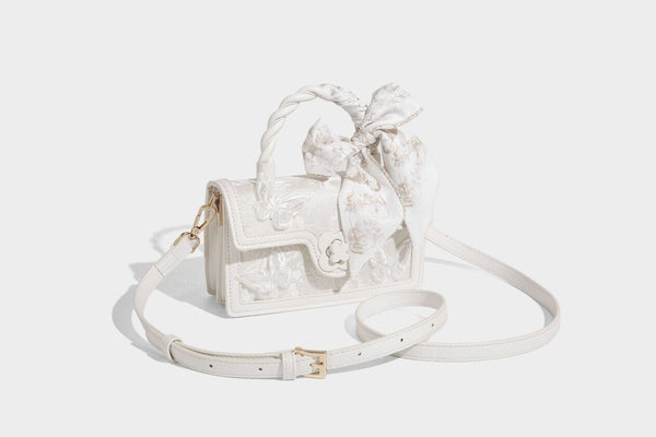 Small structured white flap bag with butterfly embroidery, flower-shaped turn-lock clasp, braided top handle with scarf bow