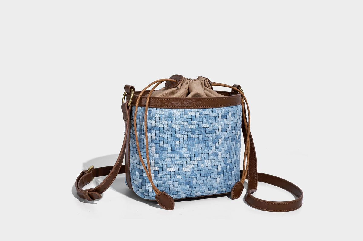 Small light blue woven bucket bag with brown leather-look trim, braided top handle, brass-tone ring hardware, drawstring