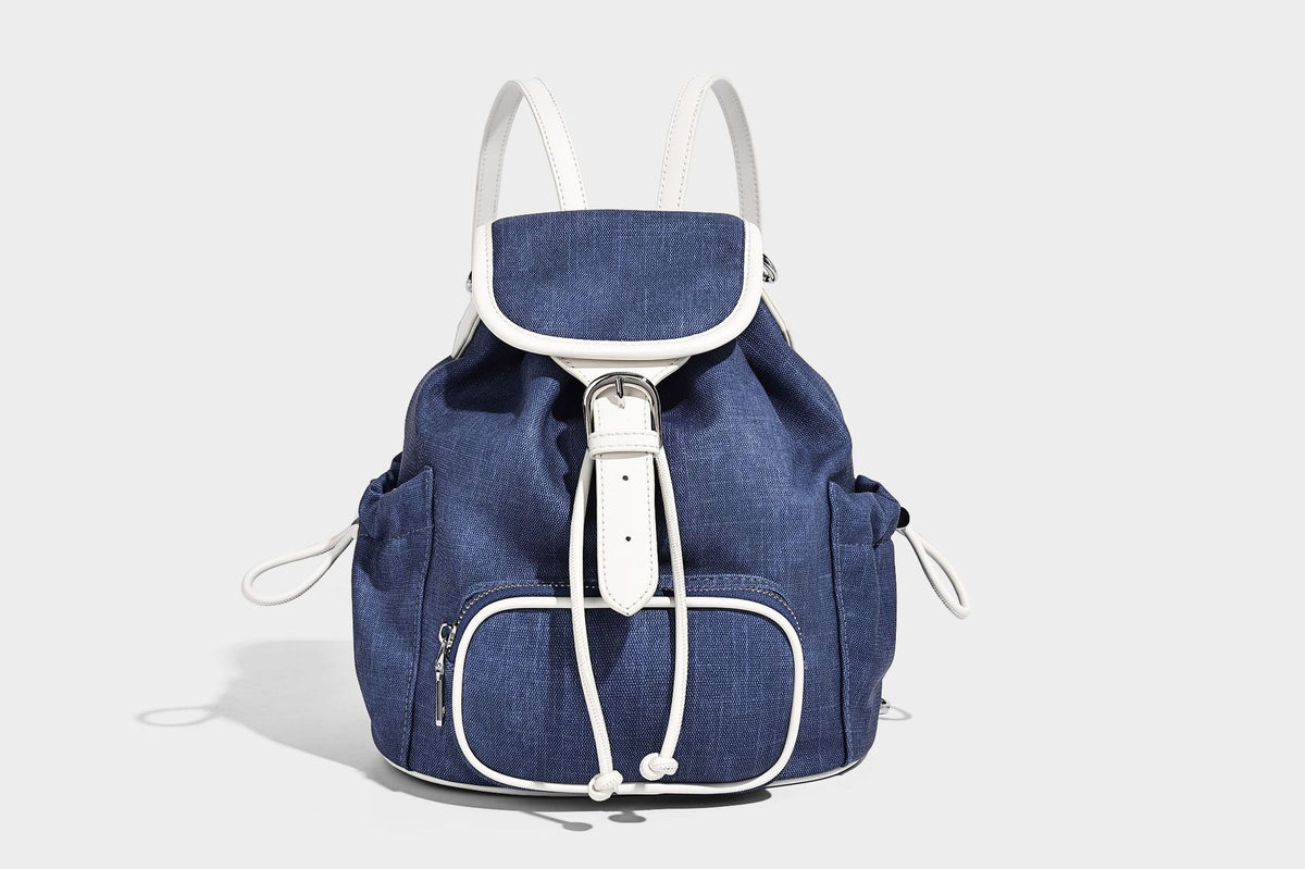 Compact denim drawstring backpack with white leather-look piping, flap-top buckle closure, front zip pocket, and silver-tone
