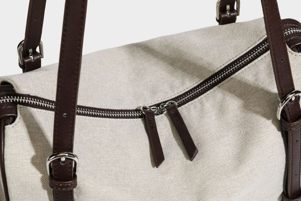Beige canvas barrel bag with dark brown leather-look trim, silver-tone hardware, top zip closure, and adjustable shoulder