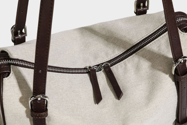 Beige canvas barrel bag with dark brown leather-look trim, silver-tone hardware, top zip closure, and adjustable shoulder