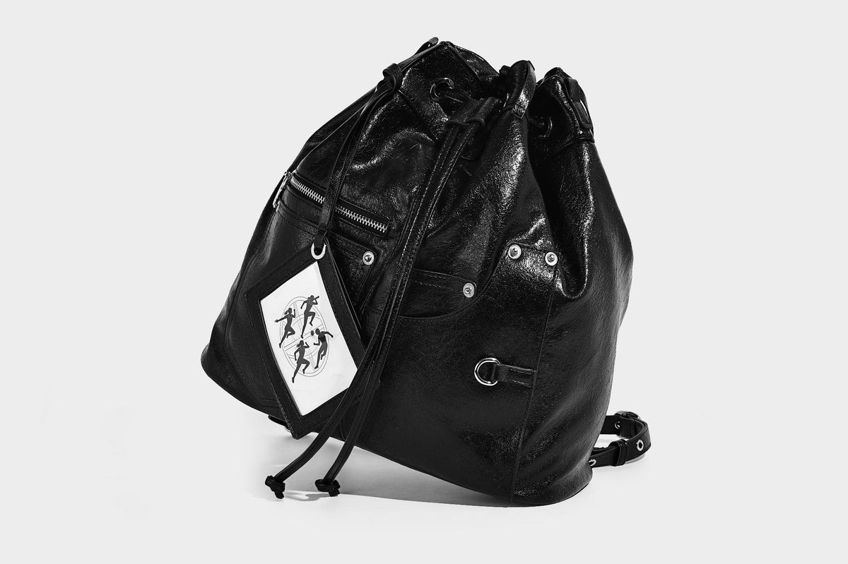 Glossy black convertible bucket backpack with silver grommet-studded straps, drawstring cinch closure, and front zip pocket