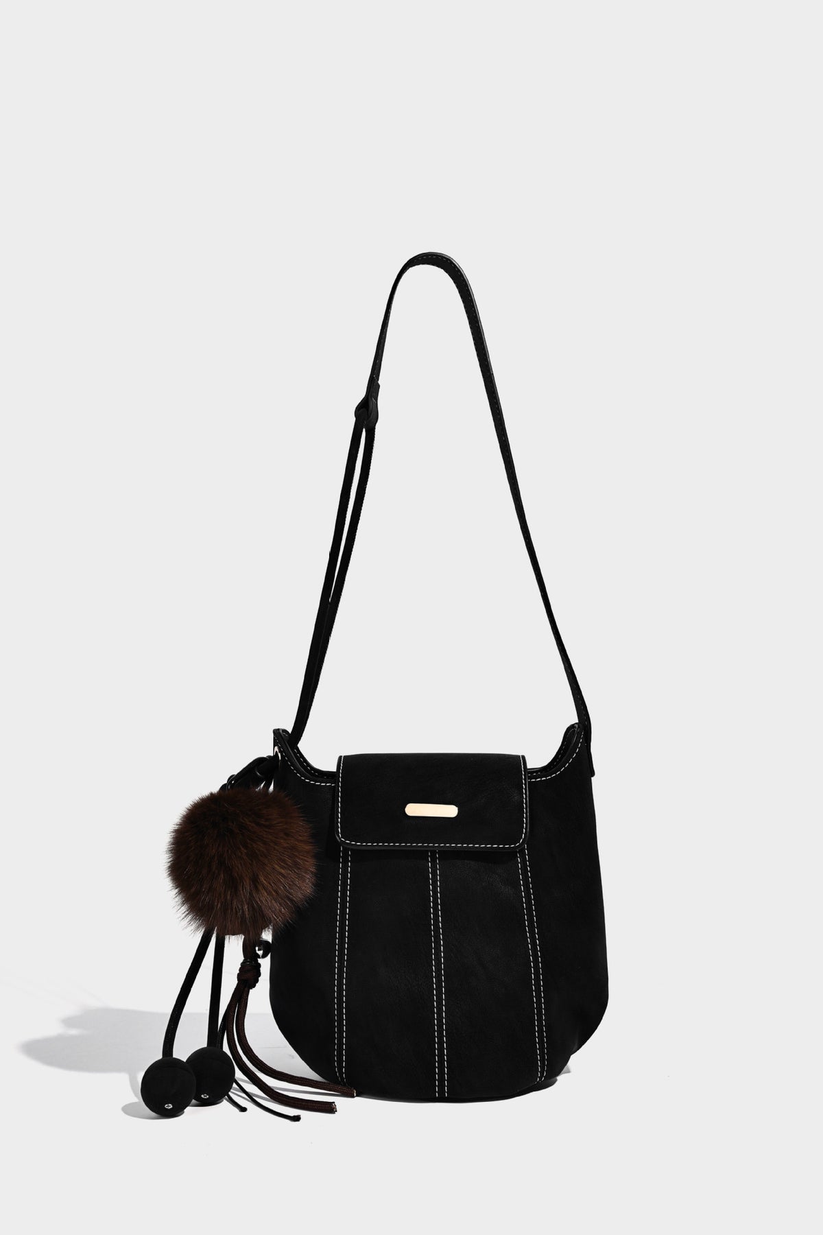 Small matte textured bucket crossbody bag with flap closure, gold-tone bar hardware, contrast stitching, and pompom tassel