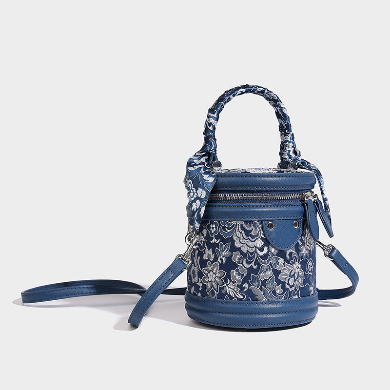 Small cylindrical barrel bag in blue-and-white floral jacquard with leather-look trim, scarf-wrapped top handle, silver-tone