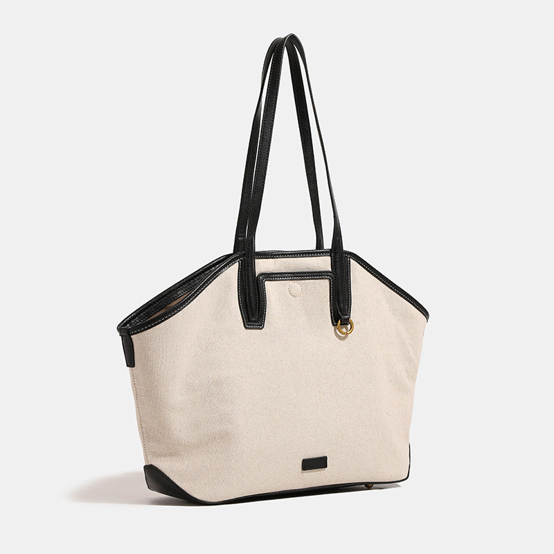 Cream canvas trapezoid tote bag with black leather-look trim, dual shoulder straps, and gold-tone ring hardware, detail view