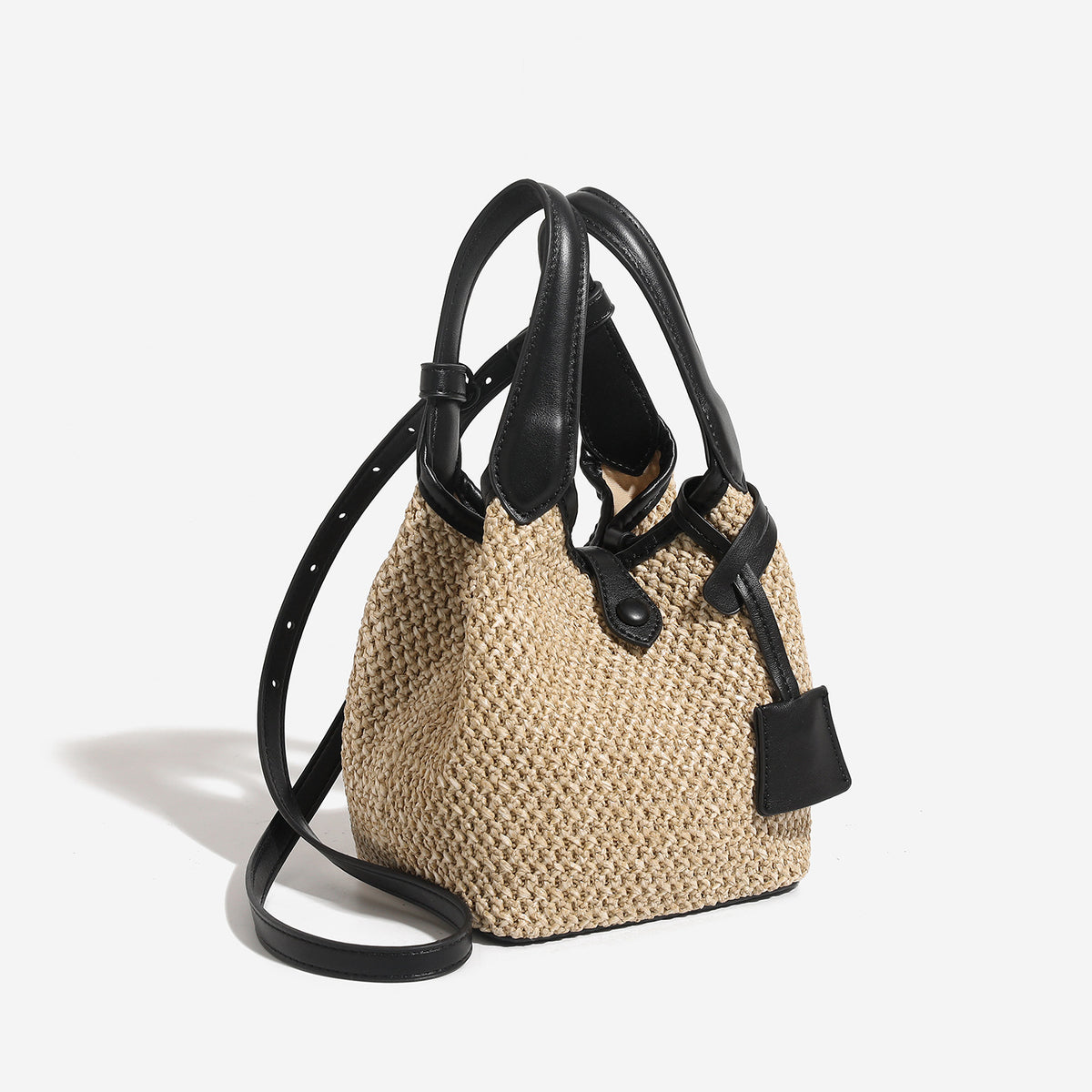 Woven beige straw bucket bag with black leather-look trim, dual top handles, detachable shoulder strap, and front snap