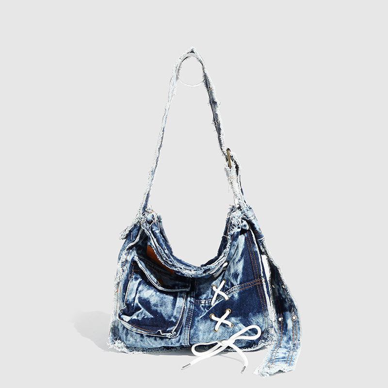 Slouchy distressed denim shoulder bag with frayed edges, white lace-up cross detail, front snap pocket, and long shoulder