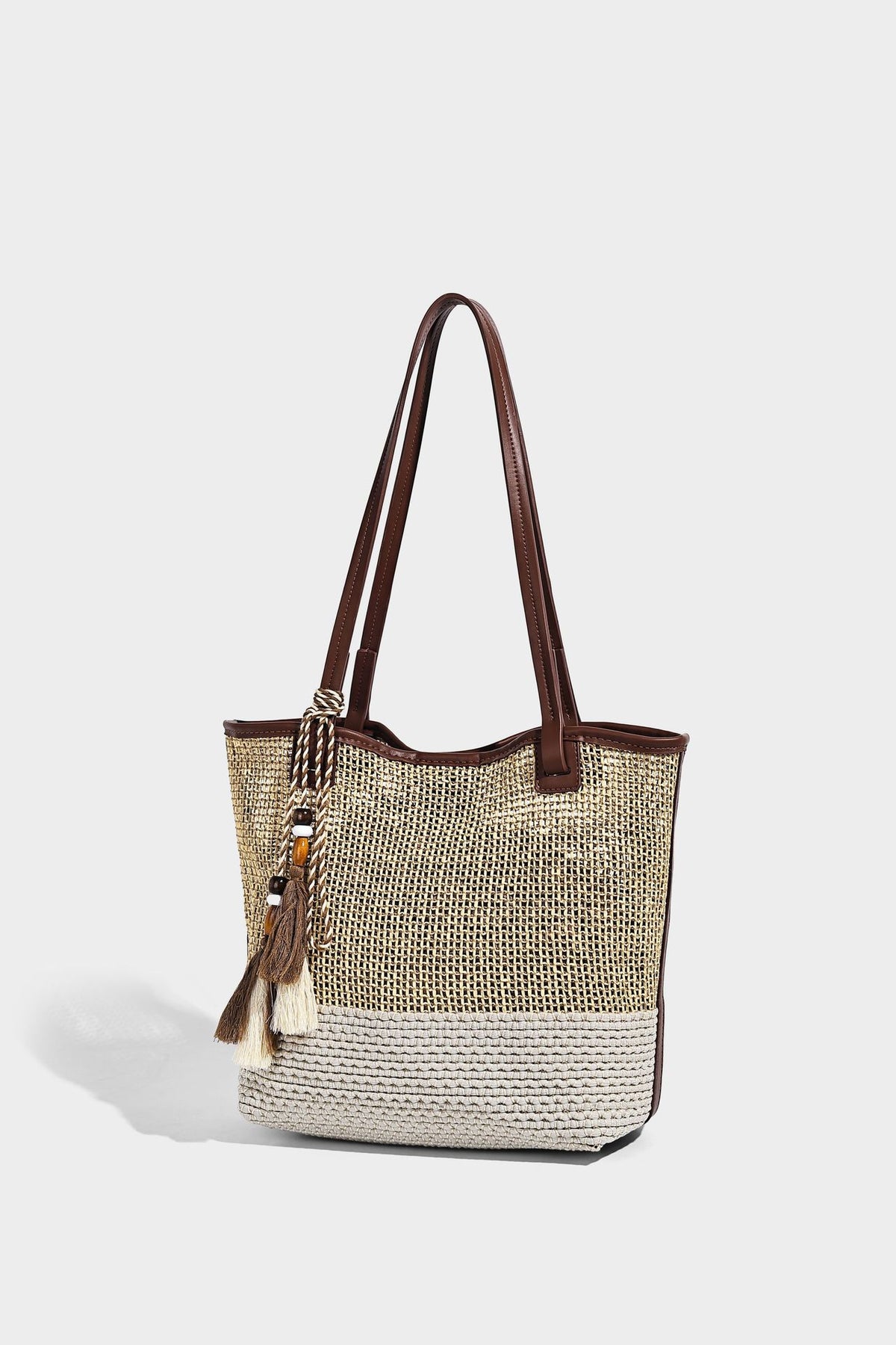 Woven beige and cream tote bag with brown leather-look trim, long double shoulder straps, and knotted tassel charm on front