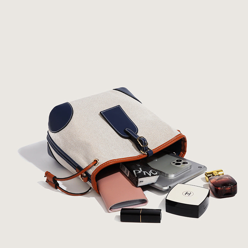 Cream woven-texture canvas bucket bag with navy accent panels, tan leather-look trim, gold-tone chain strap, and tassel