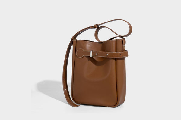 Structured dark brown leather-look bucket bag with white contrast stitching, front wraparound belt buckle strap, and long