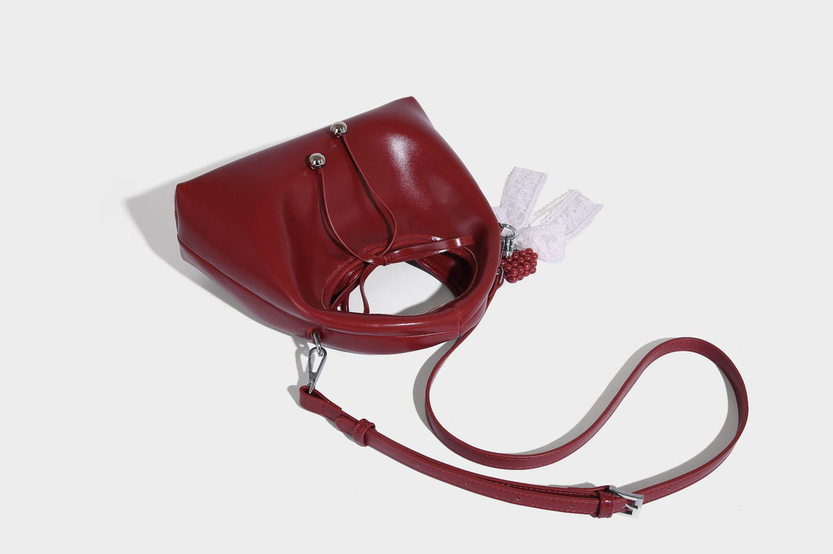 Kleine kuppelförmige Henkeltasche in glänzender Lederoptik mit Kordelzugverschluss, weißem Spitzen-Schleifenanhänger und silberfarbenen Metallbeschlägen