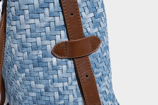 Light blue woven bucket bag with brown leather-look trim, gold-tone hardware, adjustable shoulder strap, and decorative bead
