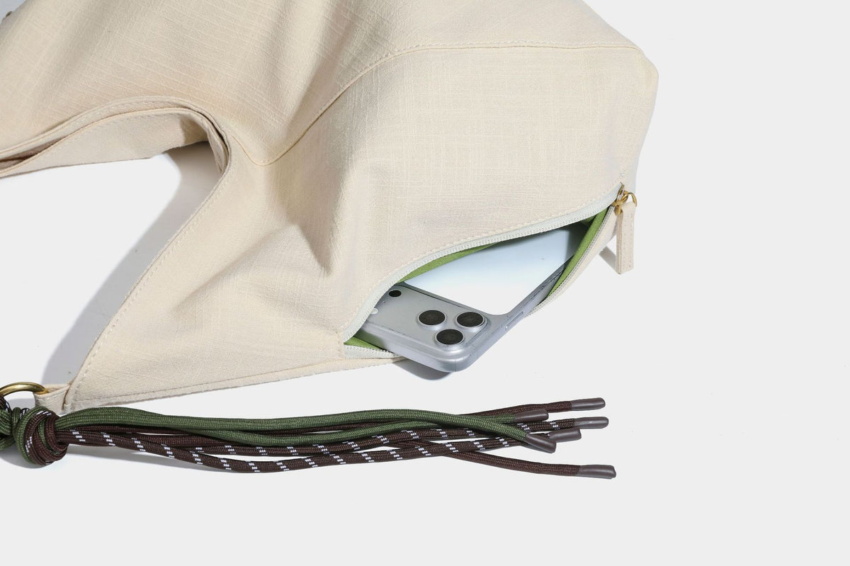 Soft slouchy cream canvas shoulder tote with braided brown and green cord handle, gold-tone ring hardware, and tassel trim
