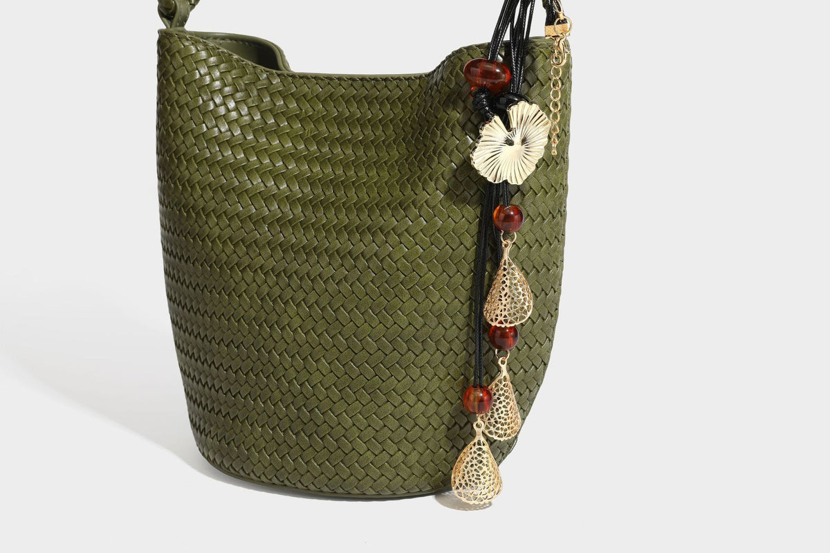 Olive green woven leather-look bucket bag with braided crossbody strap, knotted adjustment, and gold-tone tassel charm with