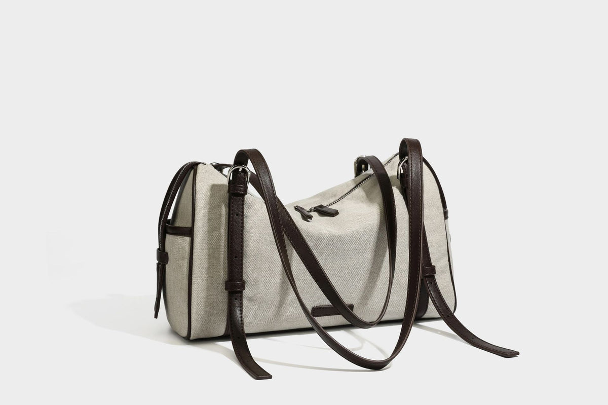 Beige canvas barrel bag with dark brown leather-look trim, silver-tone hardware, top zip closure, and adjustable shoulder