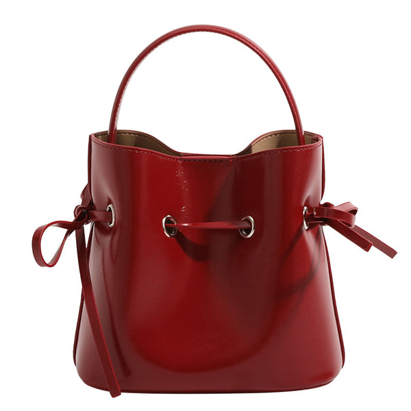 Compact glossy leather-look bucket bag with drawstring bow-tie closure, rounded top handle, and silver-tone grommet hardware