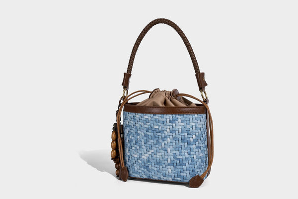 Small light blue woven bucket bag with brown leather-look trim, braided top handle, brass-tone ring hardware, drawstring