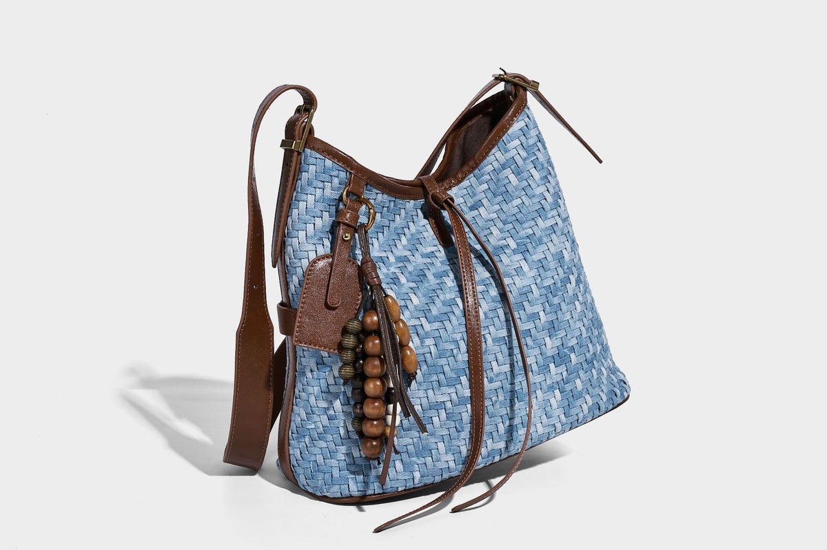 Light blue woven bucket bag with brown leather-look trim, gold-tone hardware, adjustable shoulder strap, and decorative bead