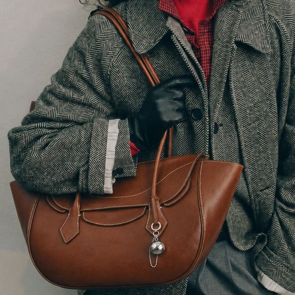 Large winged leather-look tote bag with single arched handle, stitched flap-front panel, and silver ball charm in cognac