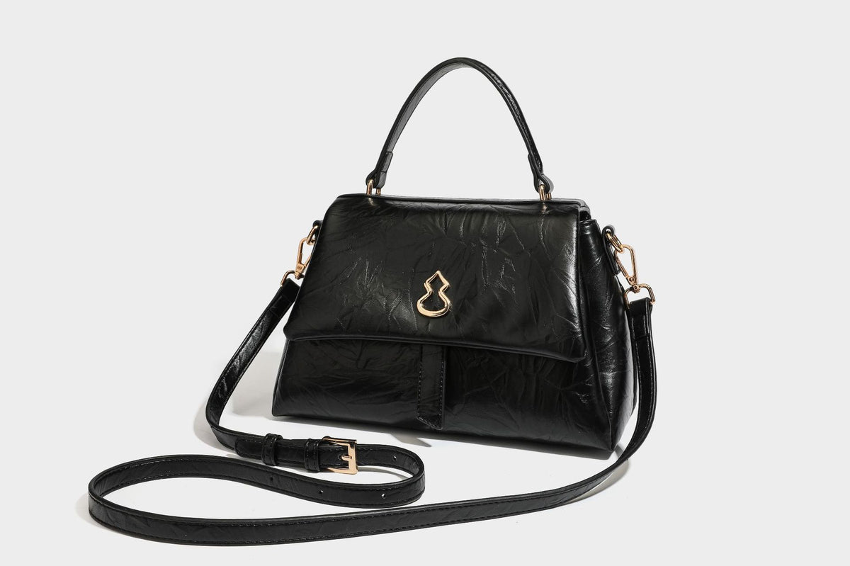 Structured black satchel bag with glossy crinkled leather-look finish, gold-tone emblem flap closure, slim top handle, and