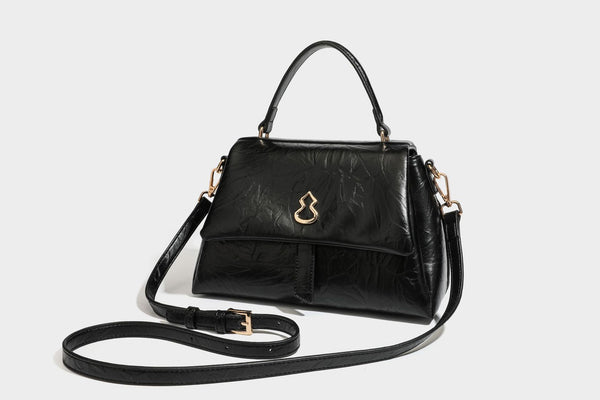 Structured black satchel bag with glossy crinkled leather-look finish, gold-tone emblem flap closure, slim top handle, and