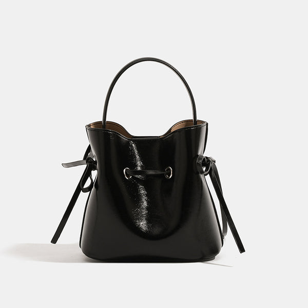 Compact glossy leather-look bucket bag with drawstring bow-tie closure, rounded top handle, and silver-tone grommet hardware