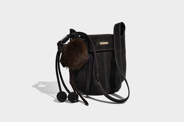 Small matte textured bucket crossbody bag with flap closure, gold-tone bar hardware, contrast stitching, and pompom tassel