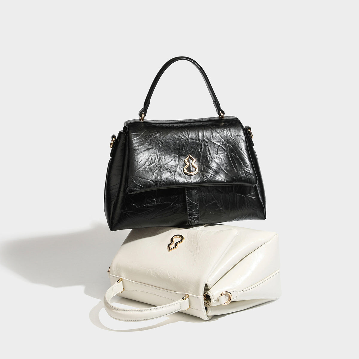 Structured black satchel bag with glossy crinkled leather-look finish, gold-tone emblem flap closure, slim top handle, and