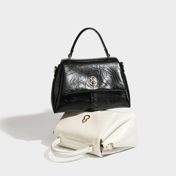 Structured black satchel bag with glossy crinkled leather-look finish, gold-tone emblem flap closure, slim top handle, and