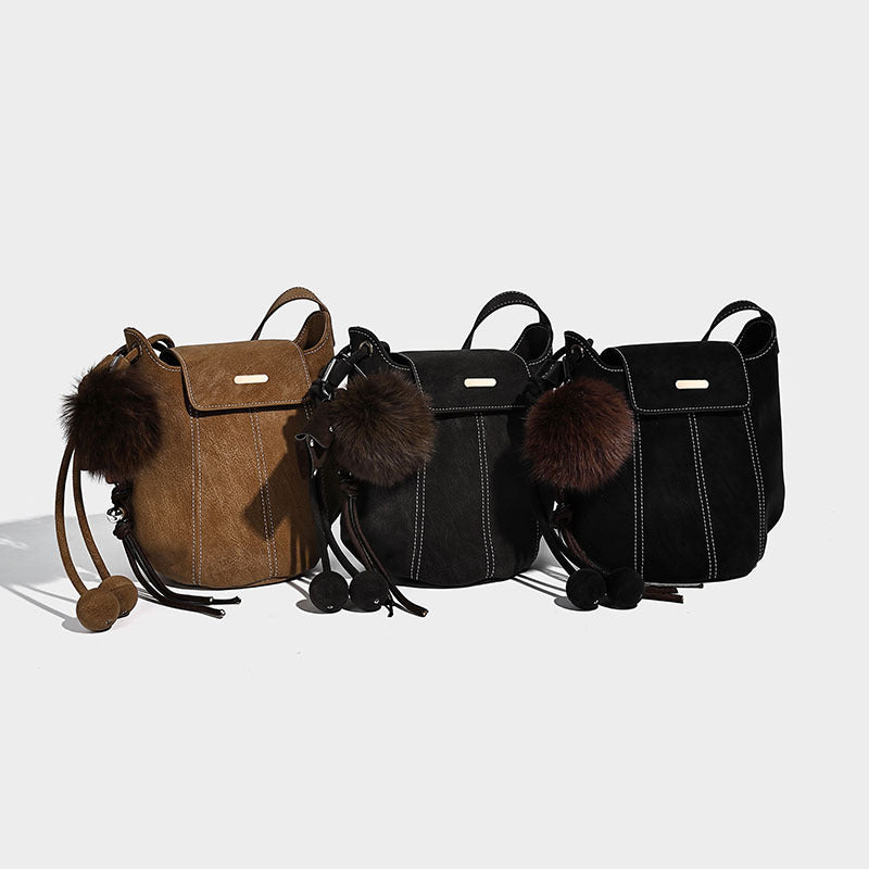 Small matte textured bucket crossbody bag with flap closure, gold-tone bar hardware, contrast stitching, and pompom tassel