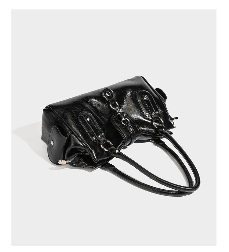 Glänzende, geknautschte Barrel-Satchel mit zwei oberen Tragegriffen, vorderem Klappen-Riemen-Panel, Ring- und Nietenbeschlägen sowie einem abnehmbaren