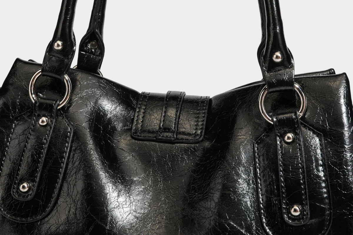 Glänzende, geknautschte Barrel-Satchel mit zwei oberen Tragegriffen, vorderem Klappen-Riemen-Panel, Ring- und Nietenbeschlägen sowie einem abnehmbaren