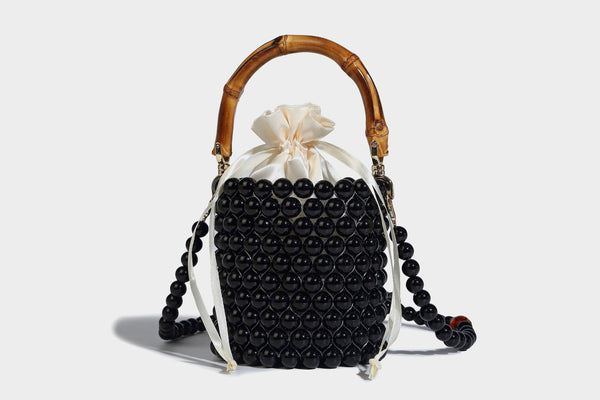 Small cylindrical bucket bag with glossy faux-pearl bead exterior, bamboo arched top handle, satin drawstring closure, and