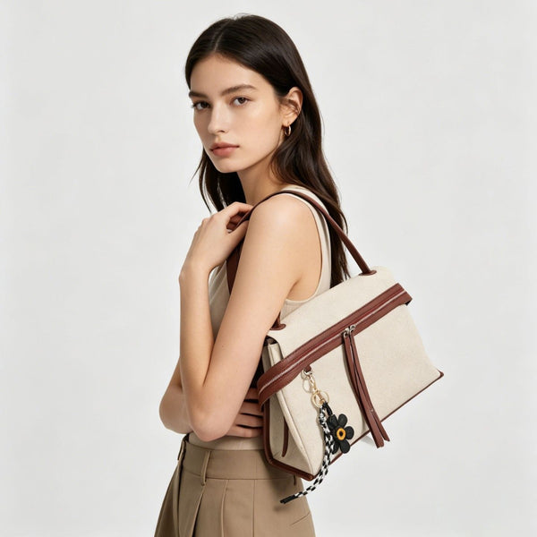 Cream canvas tote bag with brown leather-look trim, dual exterior zip pockets, long shoulder strap, and black flower charm