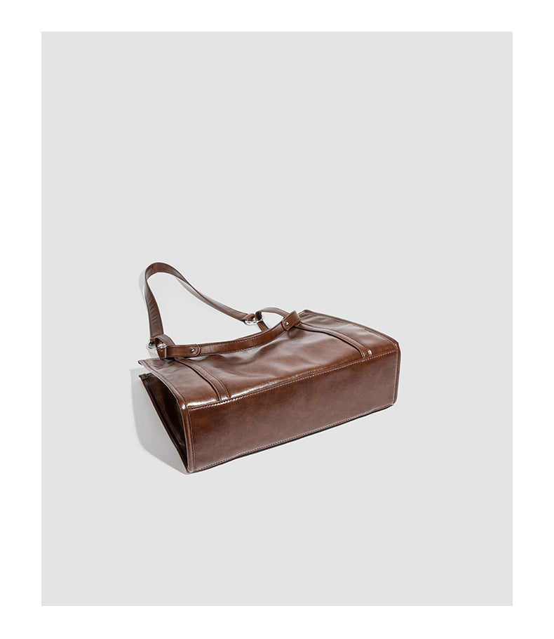 Large structured glossy leather-look tote bag with dual top handles and long shoulder strap in burnished brown, detail view