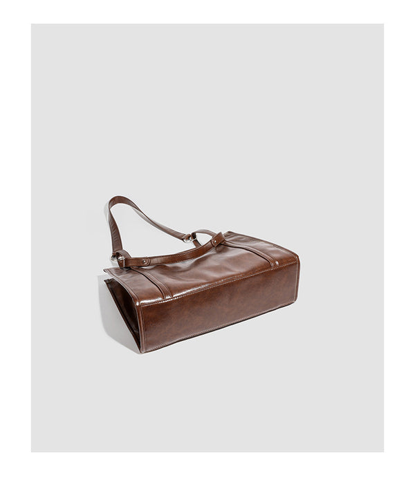 Large structured glossy leather-look tote bag with dual top handles and long shoulder strap in burnished brown, detail view