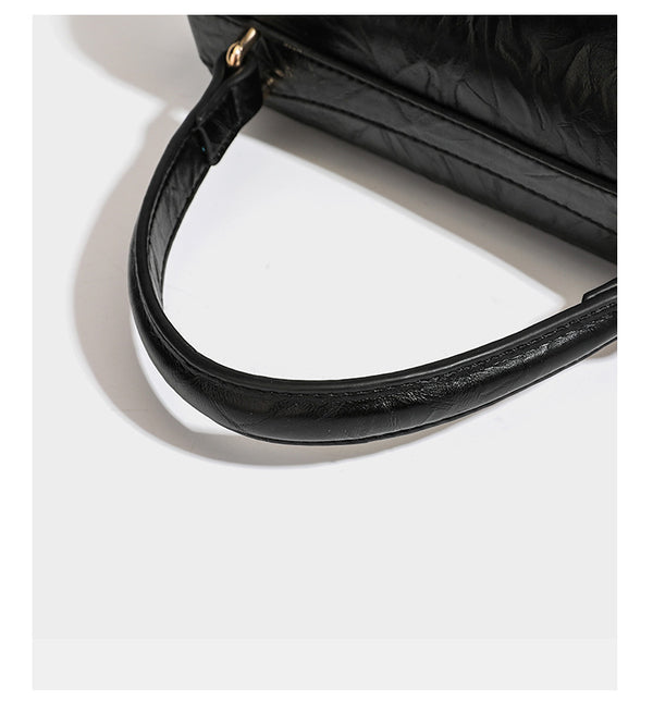 Structured black satchel bag with glossy crinkled leather-look finish, gold-tone emblem flap closure, slim top handle, and