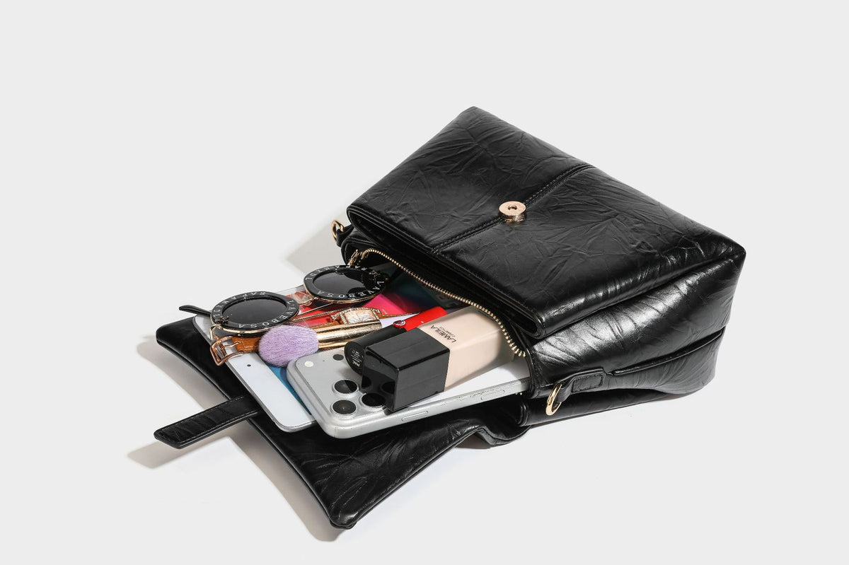 Structured black satchel bag with glossy crinkled leather-look finish, gold-tone emblem flap closure, slim top handle, and