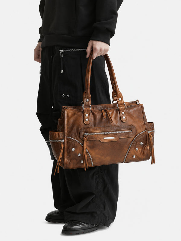 Large distressed leather-look moto tote bag with silver rivets, tassel zip pulls, front zip pocket, and dual top handles