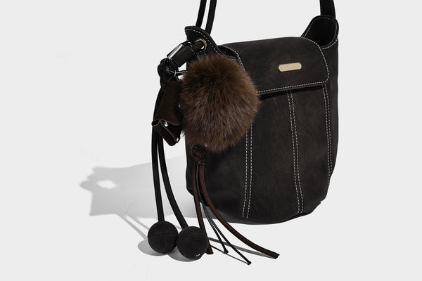Small matte textured bucket crossbody bag with flap closure, gold-tone bar hardware, contrast stitching, and pompom tassel