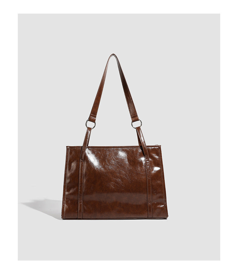 Large structured glossy leather-look tote bag with dual top handles and long shoulder strap in burnished brown, detail view
