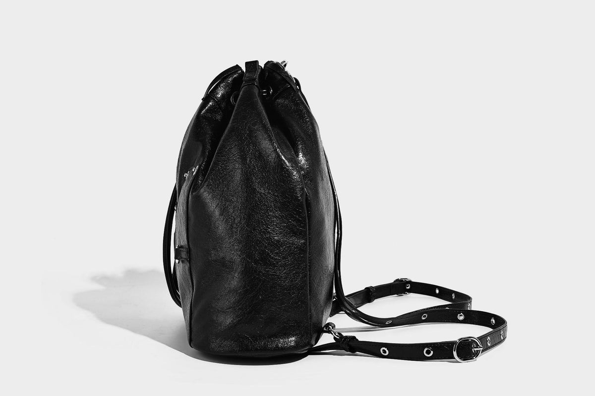 Glossy black convertible bucket backpack with silver grommet-studded straps, drawstring cinch closure, and front zip pocket