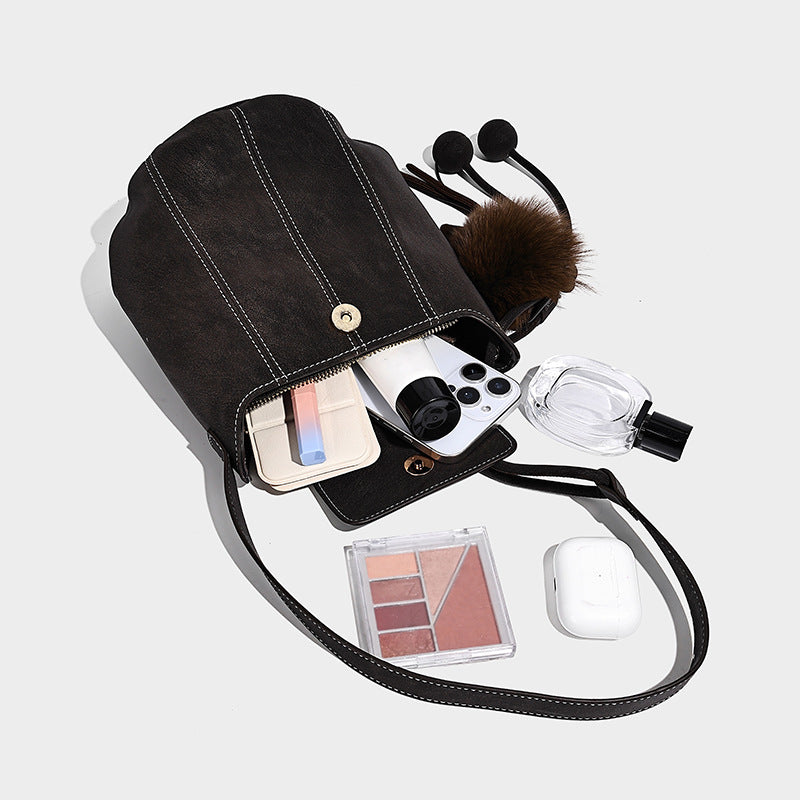 Small matte textured bucket crossbody bag with flap closure, gold-tone bar hardware, contrast stitching, and pompom tassel