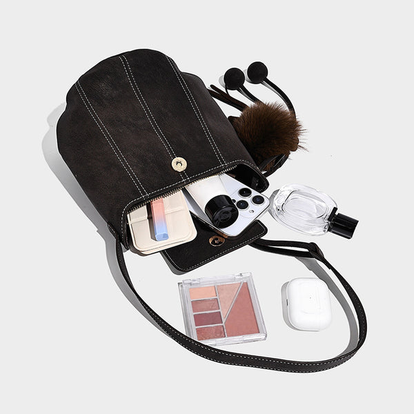 Small matte textured bucket crossbody bag with flap closure, gold-tone bar hardware, contrast stitching, and pompom tassel