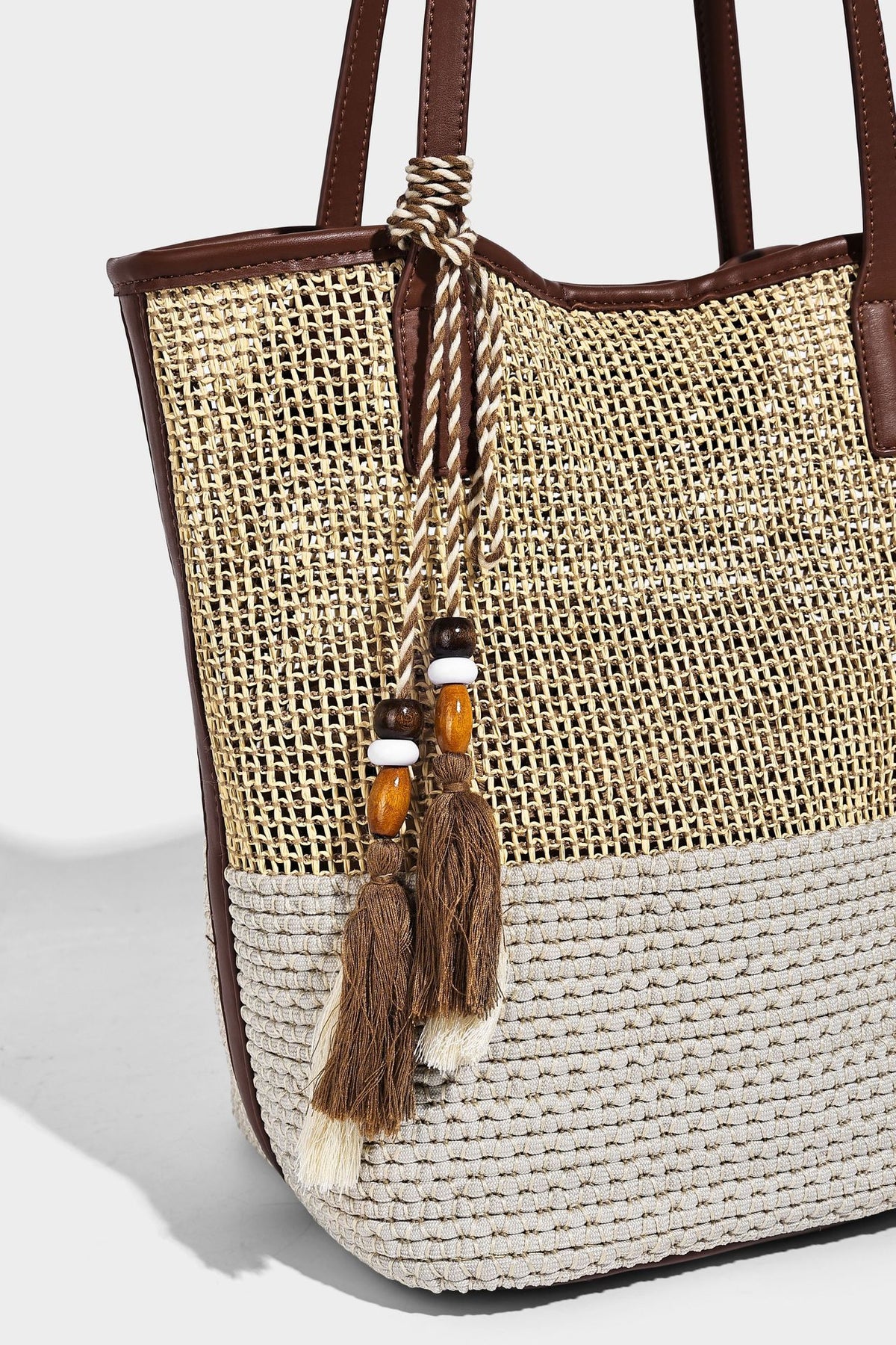 Woven straw-look tote bag in tan and cream with dark brown leather-look trim, long flat shoulder straps, and tassel bead